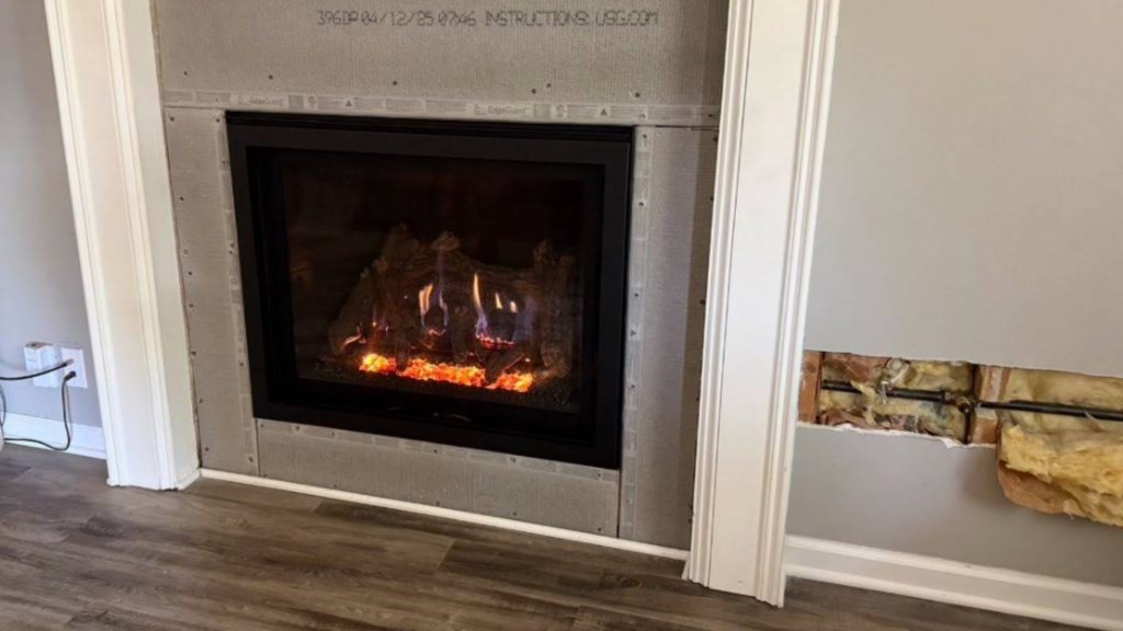 A modern fireplace glows within a framed, unfinished surround; nearby wall reveals insulation and piping